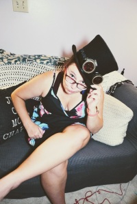 Elizabeth Parsons, a white woman with glasses, wears a top hat with goggles and sits on a couch.